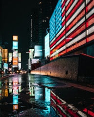 Bright neon lights and digital billboards illuminate a city street at night. A large American flag design is displayed on an electronic board, casting a colorful reflection on the wet pavement. Skyscrapers and other illuminated advertisements can be seen in the background.