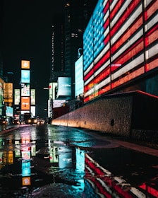 Bright neon lights and digital billboards illuminate a city street at night. A large American flag design is displayed on an electronic board, casting a colorful reflection on the wet pavement. Skyscrapers and other illuminated advertisements can be seen in the background.