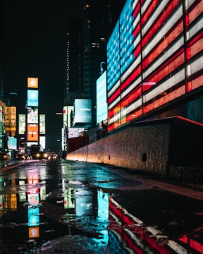 Bright neon lights and digital billboards illuminate a city street at night. A large American flag design is displayed on an electronic board, casting a colorful reflection on the wet pavement. Skyscrapers and other illuminated advertisements can be seen in the background.
