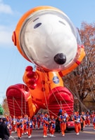 A large inflatable balloon of a cartoon dog dressed in an astronaut suit, accompanied by handlers in matching orange costumes walking alongside. The balloon features a helmet with a clear visor, large red boots, and an orange suit with a NASA logo. Trees with autumn foliage and a clear blue sky form the background.