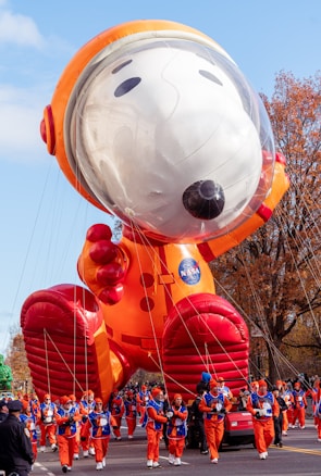 A large inflatable balloon of a cartoon dog dressed in an astronaut suit, accompanied by handlers in matching orange costumes walking alongside. The balloon features a helmet with a clear visor, large red boots, and an orange suit with a NASA logo. Trees with autumn foliage and a clear blue sky form the background.