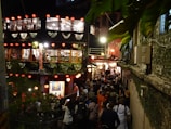 The scene depicts a lively, bustling street at night with a crowd of people gathered in front of a two-story building adorned with red lanterns and green plants. The building has large windows showing a brightly lit interior. The atmosphere appears festive and warm, enhanced by the lighting and decorative elements. The setting is likely a popular tourist spot or cultural area.