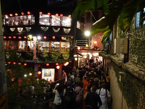 The scene depicts a lively, bustling street at night with a crowd of people gathered in front of a two-story building adorned with red lanterns and green plants. The building has large windows showing a brightly lit interior. The atmosphere appears festive and warm, enhanced by the lighting and decorative elements. The setting is likely a popular tourist spot or cultural area.
