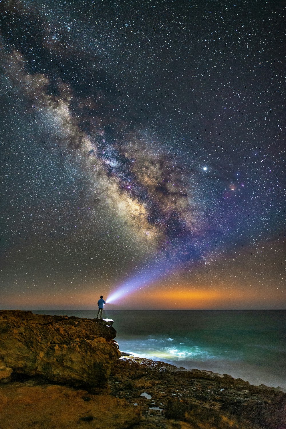 person standing near cliff holding flashlight overlooking milky way