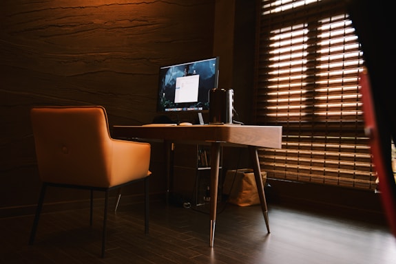 A modern office space featuring a wooden desk with a large monitor and a sleek computer setup. An orange upholstered chair is positioned in front of the desk. There is soft lighting and a window with horizontal blinds, casting a warm ambiance.