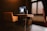 A modern office space featuring a wooden desk with a large monitor and a sleek computer setup. An orange upholstered chair is positioned in front of the desk. There is soft lighting and a window with horizontal blinds, casting a warm ambiance.