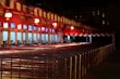 A row of ticket booths under a red and white striped canopy is lit with warm lights. The booths display digital screens and are arranged along metal railings. The scene is illuminated in a night setting with an empty path leading towards the booths.
