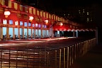 A row of ticket booths under a red and white striped canopy is lit with warm lights. The booths display digital screens and are arranged along metal railings. The scene is illuminated in a night setting with an empty path leading towards the booths.