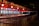A row of ticket booths under a red and white striped canopy is lit with warm lights. The booths display digital screens and are arranged along metal railings. The scene is illuminated in a night setting with an empty path leading towards the booths.