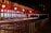 A row of ticket booths under a red and white striped canopy is lit with warm lights. The booths display digital screens and are arranged along metal railings. The scene is illuminated in a night setting with an empty path leading towards the booths.
