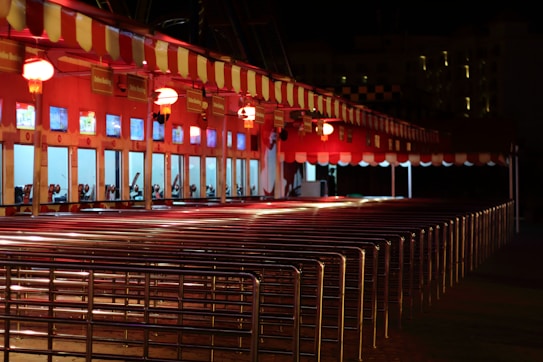 A row of ticket booths under a red and white striped canopy is lit with warm lights. The booths display digital screens and are arranged along metal railings. The scene is illuminated in a night setting with an empty path leading towards the booths.