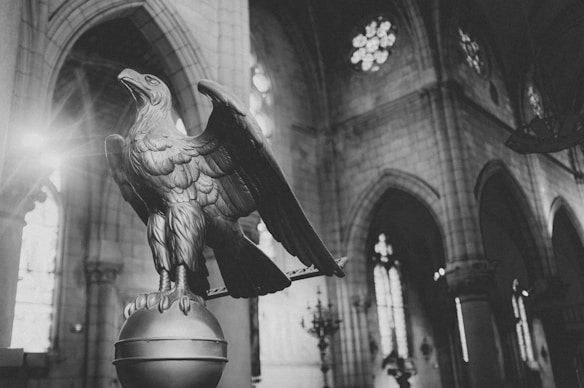 A black and white photograph of a cathedral interior featuring a prominent eagle sculpture perched on a globe. The architecture includes tall, arched windows and intricate stonework with visible stained glass details. Soft natural light filters through the windows creating a serene atmosphere.