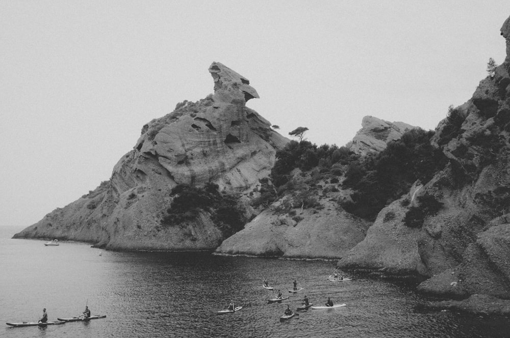grayscale photo of group of people on clam body of water in front of rock formation
