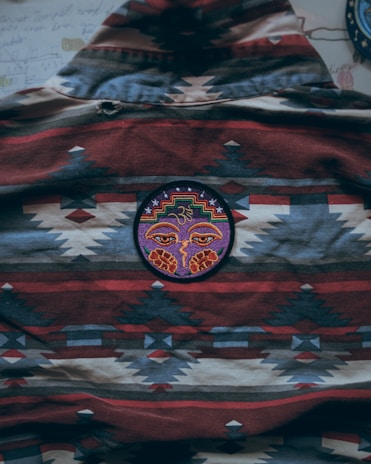 A piece of fabric with a geometric and tribal pattern featuring a mixture of red, blue, beige, and black. In the center, there is a circular patch with an intricate design of a face adorned with colorful patterns, including a symbol resembling a spiral or third eye.