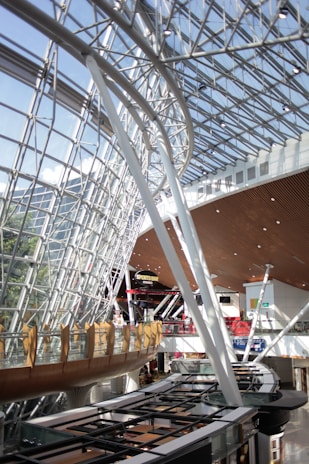 A spacious and modern interior of a building with a large glass and steel ceiling, featuring curved lines and a wooden walkway. The architecture allows natural light to flood the area, creating a bright and airy atmosphere. A sports bar with signage is visible, along with escalators and various retail spaces beneath the walkway.