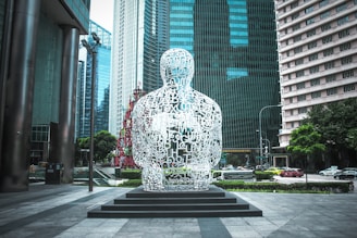 A large sculpture made of interconnected white letters forms the shape of a seated human figure. It is situated on a pedestal in an urban area, surrounded by modern skyscrapers and a city street with cars. Trees and greenery add contrast to the urban environment.