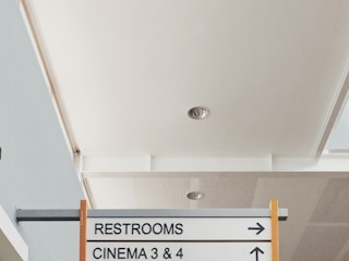 A directional sign in an indoor setting, pointing to restrooms and cinema 3 & 4. The ceiling above is smoothly finished with recessed lighting.