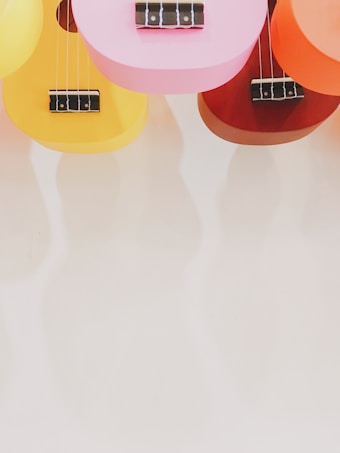 Several colorful ukuleles are arranged with their bodies facing downward. They are in vibrant shades of pink, yellow, orange, and red, and their black bridges and white strings are visible. The clean, light background enhances the brightness of the ukuleles.