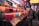 A vibrant array of colorful drinks is displayed in plastic cups at a busy market stall. Signs with prices and drink names are arranged in front of each row. Customers stand in front of the stall, browsing the options.