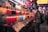 A vibrant array of colorful drinks is displayed in plastic cups at a busy market stall. Signs with prices and drink names are arranged in front of each row. Customers stand in front of the stall, browsing the options.