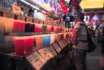 A vibrant array of colorful drinks is displayed in plastic cups at a busy market stall. Signs with prices and drink names are arranged in front of each row. Customers stand in front of the stall, browsing the options.