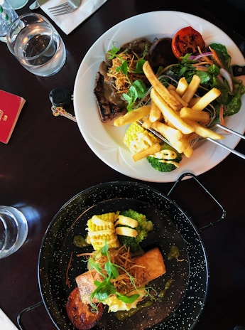 Two plates of gourmet food are placed on a dark table. The top plate features a steak topped with a rich brown sauce, accompanied by french fries, corn on the cob, grilled tomato, and a fresh salad. The bottom plate presents a piece of salmon with a creamy sauce, broccoli, more grilled tomato, and garnished with herbs. Beside the plates are two glasses of water and a red notebook with a pen.