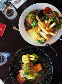 Two plates of gourmet food are placed on a dark table. The top plate features a steak topped with a rich brown sauce, accompanied by french fries, corn on the cob, grilled tomato, and a fresh salad. The bottom plate presents a piece of salmon with a creamy sauce, broccoli, more grilled tomato, and garnished with herbs. Beside the plates are two glasses of water and a red notebook with a pen.