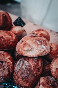 A collection of rustic, artisanal bread loaves stacked on a wooden surface, with a small blackboard sign in the background. The loaves have a crusty, textured surface dusted with flour, showcasing a rich brown color with lighter patches.