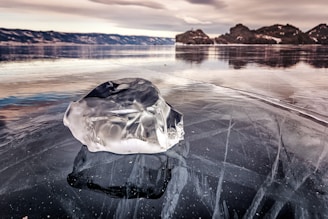 A clear, smooth piece of ice rests on the surface of a frozen lake, with visible cracks and patterns beneath it. The background features snow-capped mountains and a cloudy sky, creating a tranquil and cold atmosphere.