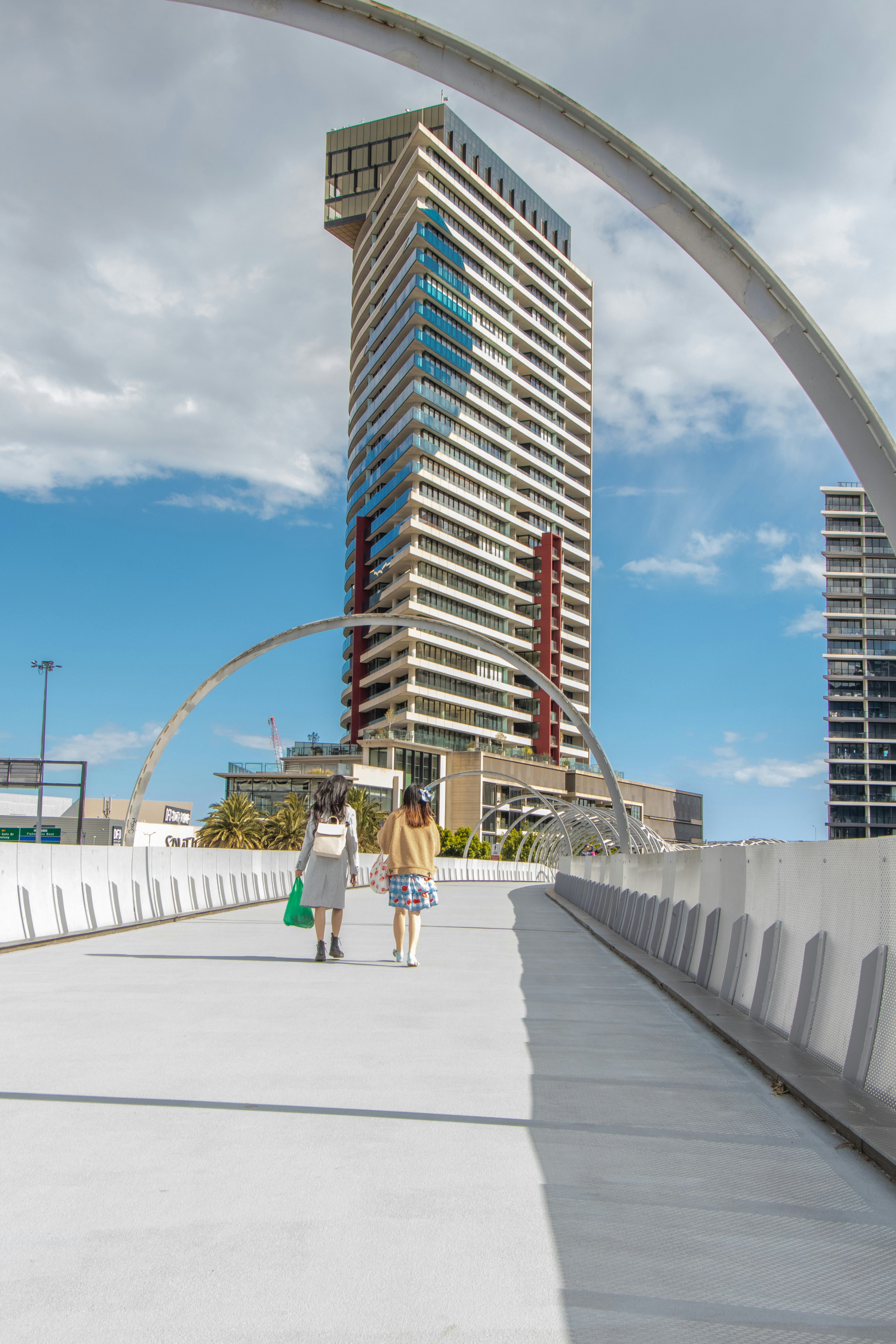 Two pedestrians walk along a bright elevated walkway toward a tall, modern tower framed by a sweeping arch on a sunny urban day.