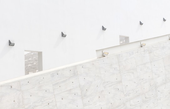 A minimalist architectural scene featuring a white wall with geometric shapes and modern wall-mounted lights. The wall has several square openings and reflects a play of light and shadow, creating a clean and contemporary aesthetic.