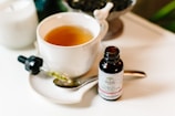 A white ceramic cup filled with a light brown liquid is placed on a matching saucer. Beside the cup, there is a spoon and a small bottle labeled 'hemp oil CBD drops,' along with a dropper filled with a yellowish liquid. The background includes a white candle and some greenery.