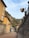 Narrow street lined with buildings featuring rustic, warm-toned facades. A sign for Hotel Villa Cipressi is prominently displayed on the right. Lush green hills with tall, slender trees rise in the background under a partly cloudy sky.