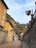 Narrow street lined with buildings featuring rustic, warm-toned facades. A sign for Hotel Villa Cipressi is prominently displayed on the right. Lush green hills with tall, slender trees rise in the background under a partly cloudy sky.