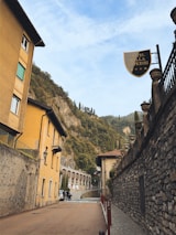 Narrow street lined with buildings featuring rustic, warm-toned facades. A sign for Hotel Villa Cipressi is prominently displayed on the right. Lush green hills with tall, slender trees rise in the background under a partly cloudy sky.