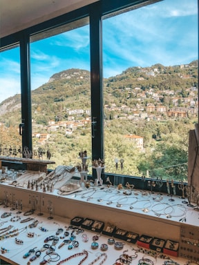 A scenic window view overlooks a hilly landscape dotted with houses and lush greenery. In the foreground, a collection of jewelry, including necklaces, earrings, and rings, is neatly arranged on a table.