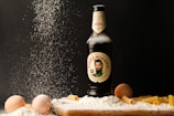 A bottle of Birra Moretti is surrounded by a scattering of flour, eggs, and uncooked rigatoni pasta. The flour appears to be falling from above, creating a dynamic effect against the dark background. The bottle is upright and partially dusted with flour.