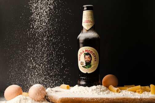A bottle of Birra Moretti is surrounded by a scattering of flour, eggs, and uncooked rigatoni pasta. The flour appears to be falling from above, creating a dynamic effect against the dark background. The bottle is upright and partially dusted with flour.