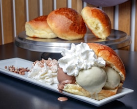 A delicious plate featuring a dessert consisting of a brioche bun filled with scoops of chocolate and vanilla ice cream, topped with whipped cream. The dessert is served on a rectangular white plate next to additional whipped cream garnished with cocoa powder and small chocolate pearls. In the background, two plain brioche buns are displayed on a metal tray.