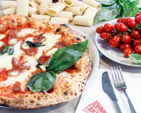 A delicious-looking pizza with a bubbly crust, topped with melted cheese, pepperoni, and fresh basil leaves. Surrounding the pizza are uncooked pasta tubes, a plate of vibrant red cherry tomatoes, and fresh basil leaves. A fork and knife are placed on a napkin nearby, with the text 'JOHNNY TAKE(S)' visible on it.