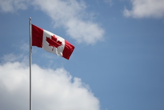 A Canadian flag is flying on a flagpole against a backdrop of a blue sky with scattered clouds. The flag displays a red maple leaf in the center, with red bars on either side.