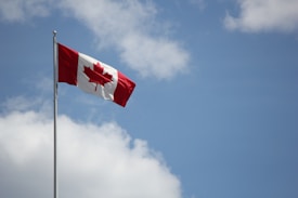 A Canadian flag is flying on a flagpole against a backdrop of a blue sky with scattered clouds. The flag displays a red maple leaf in the center, with red bars on either side.