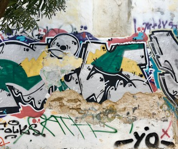 A vibrant piece of street art featuring abstract shapes and patterns. The wall is partially deteriorated, adding an urban, gritty texture. Green, yellow, black, and red colors stand out against the white background. Graffiti tags and various symbols are visible, with portions of peeling paint and exposed concrete.