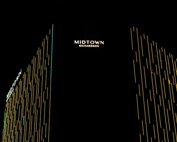 Vertical lines of yellow light create a modern, sleek design on a high-rise building. The name 'Midtown Richardson' is prominently displayed in illuminated text at the top, with the structure captured against a dark night sky.