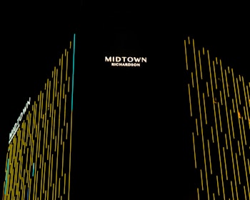Vertical lines of yellow light create a modern, sleek design on a high-rise building. The name 'Midtown Richardson' is prominently displayed in illuminated text at the top, with the structure captured against a dark night sky.