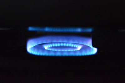 A blue flame from a gas stove burner is prominently displayed against a dark background. The flame forms a circular pattern with a soft, glowing appearance.