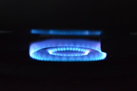 A blue flame from a gas stove burner is prominently displayed against a dark background. The flame forms a circular pattern with a soft, glowing appearance.