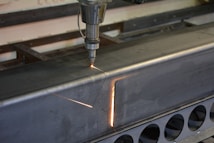 A metalworking machine is in operation, cutting a rectangular shape into a large metal beam. Sparks and a bright cutting line indicate the active laser or cutting tool. The setting appears to be a workshop or industrial environment with visible metal fixtures and structures.