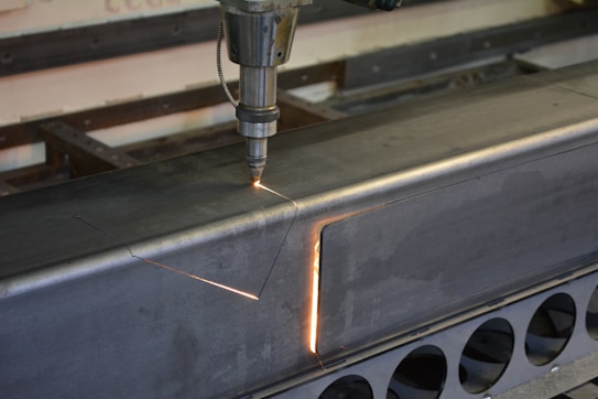 A metalworking machine is in operation, cutting a rectangular shape into a large metal beam. Sparks and a bright cutting line indicate the active laser or cutting tool. The setting appears to be a workshop or industrial environment with visible metal fixtures and structures.
