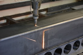 A metalworking machine is in operation, cutting a rectangular shape into a large metal beam. Sparks and a bright cutting line indicate the active laser or cutting tool. The setting appears to be a workshop or industrial environment with visible metal fixtures and structures.